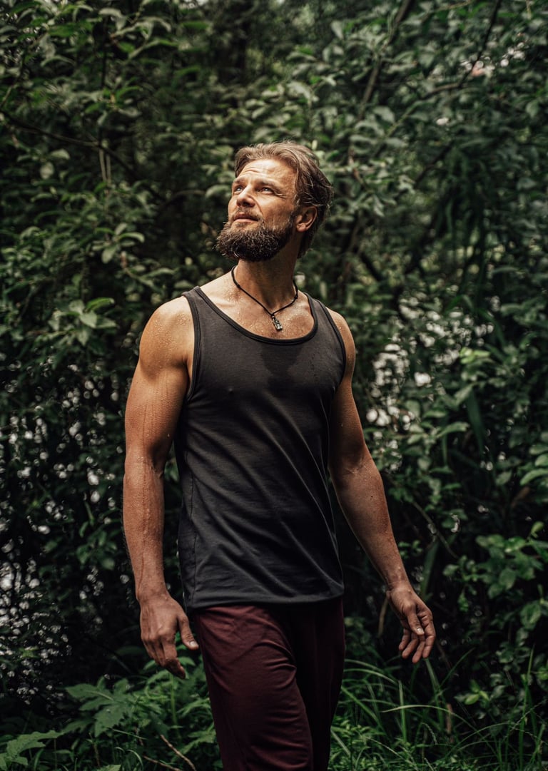 Bearded man in black tank top and burgundy pants standing in front of lush green foliage, gazing upward